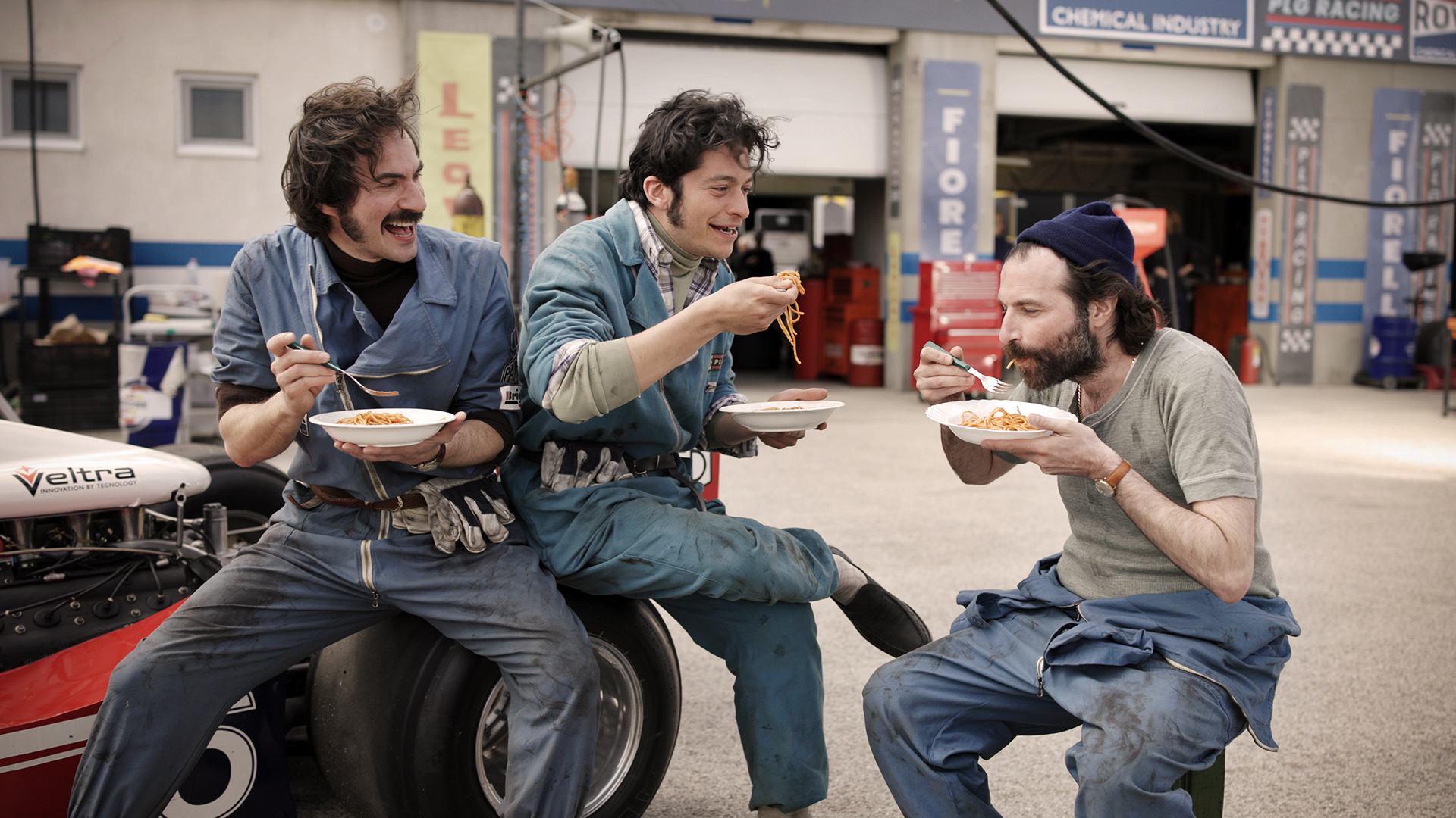 Period race mechanics share a plate of spaghetti in the pit lane — a scene from the film.