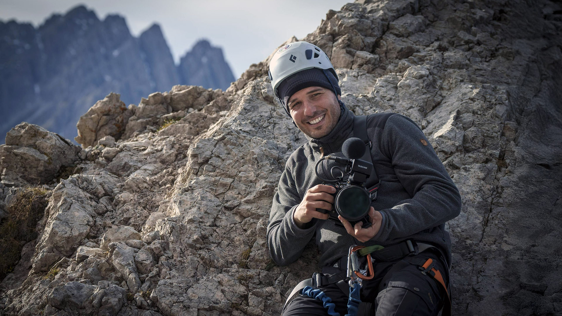 Filming on a rock face in a climbing helmet, camera and shotgun mic in hand.