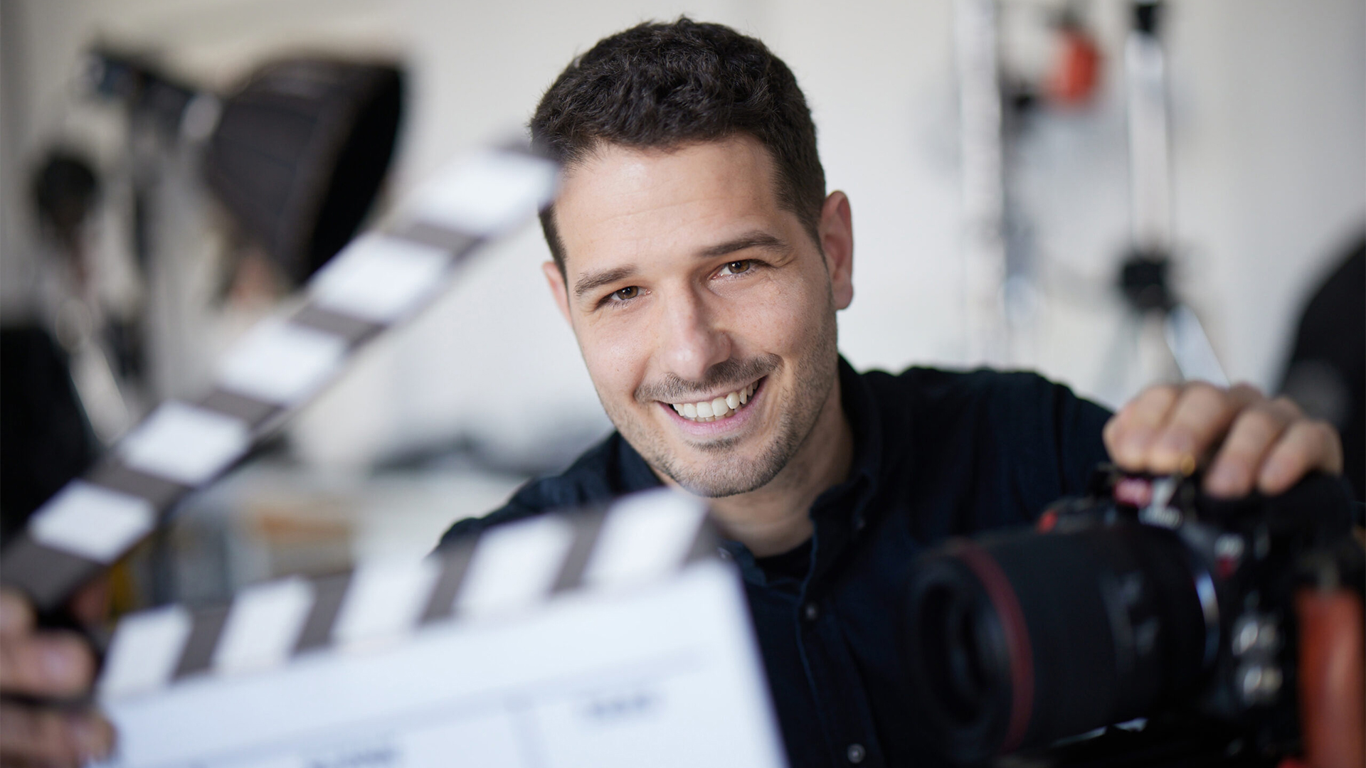 Portrait of András Biró smiling behind a cinema camera, clapperboard in hand.