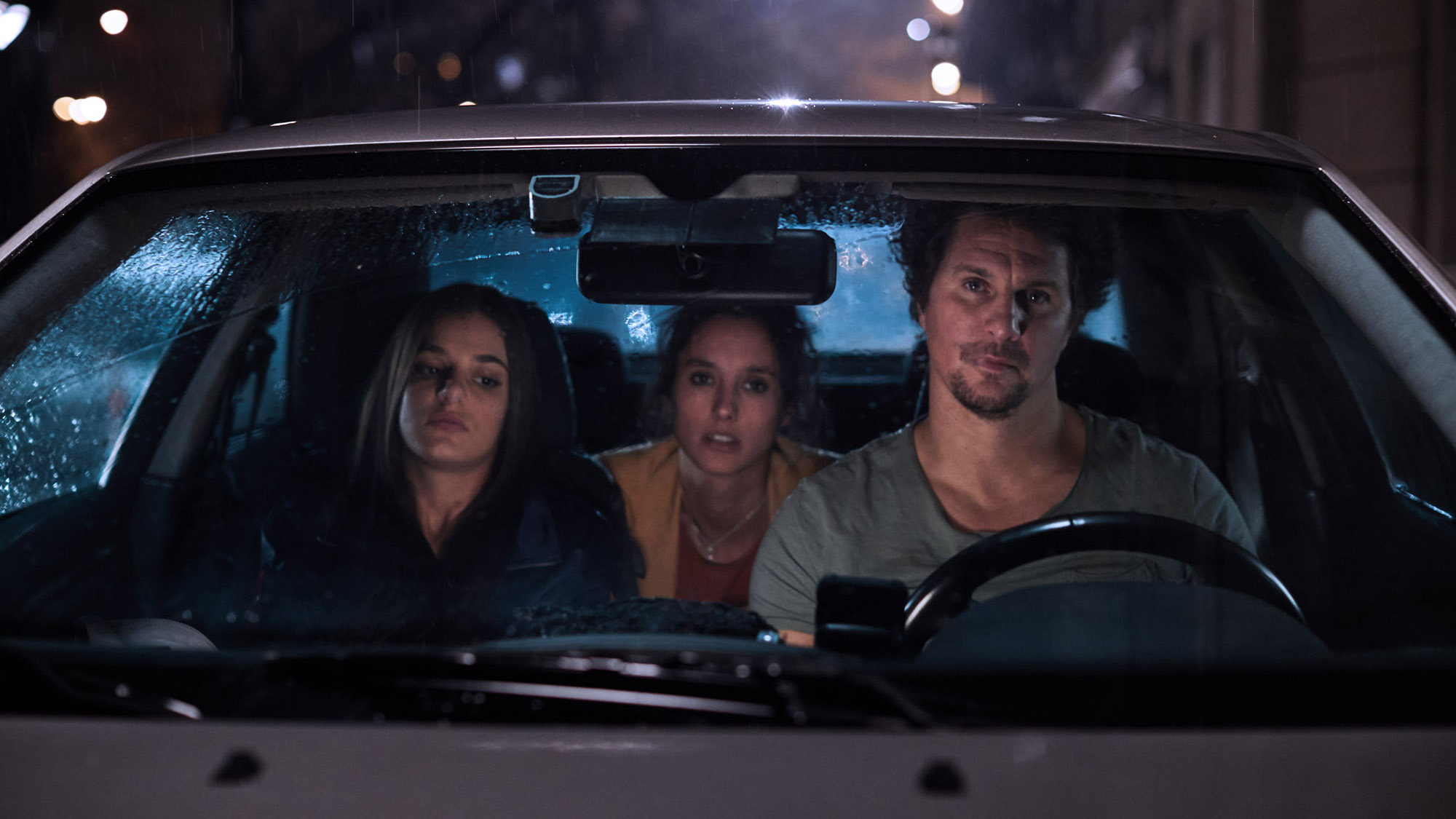 Night scene through a rain-soaked windscreen: three passengers in a parked car.