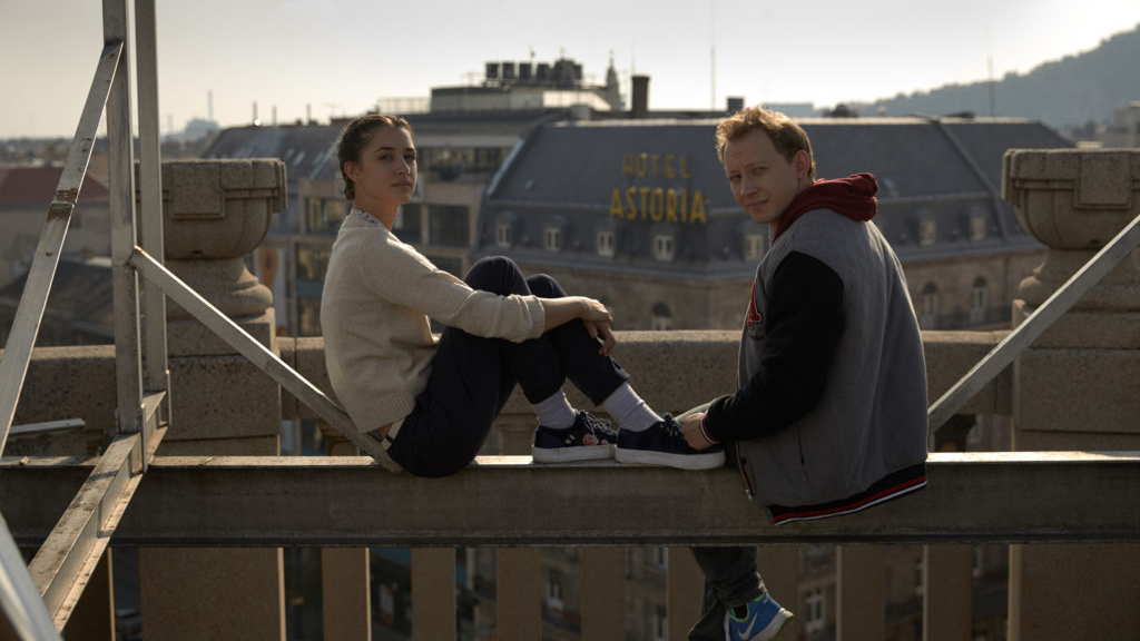 Two actors on a rooftop ledge above the city, the Hotel Astoria behind them.